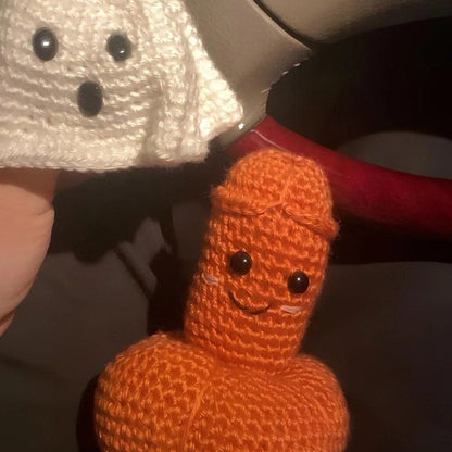 Close-up of a cheeky orange crochet penis-shaped Halloween novelty decoration with a smiling face, displayed beside a white ghost.